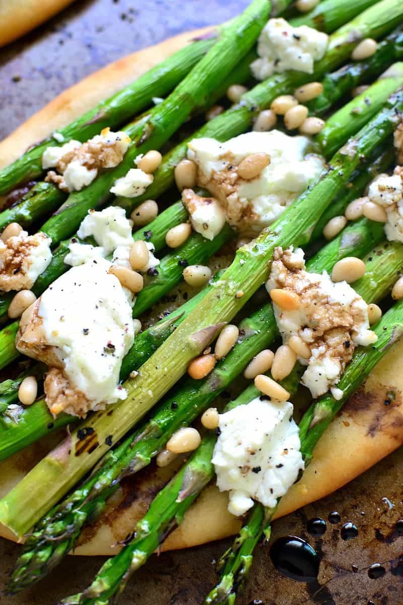 close up image of goat cheese melting over the top of asparagus and pine nuts on flatbread