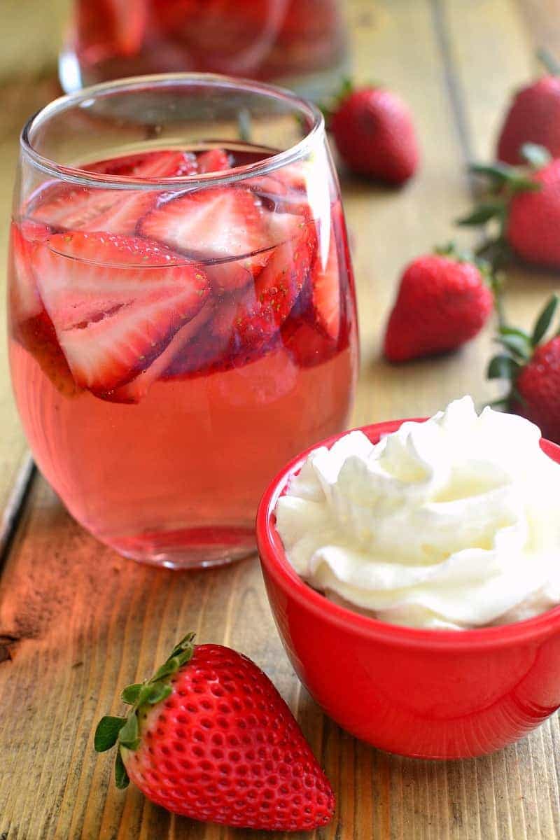 a glass of moscato wine sangria with fresh strawberry slices - glass is surrounded by whole strawberries and a bowl of whipped cream