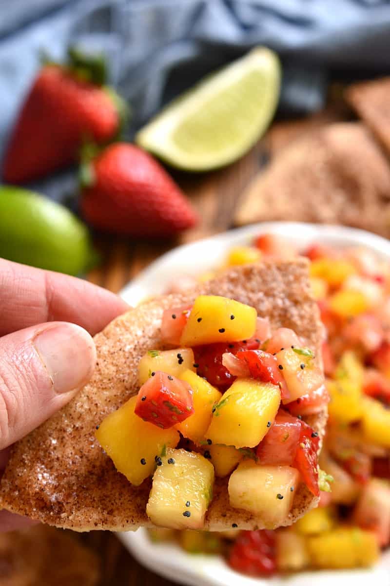 person holding a tortilla chip, dipping into a bowl of fruit salsa