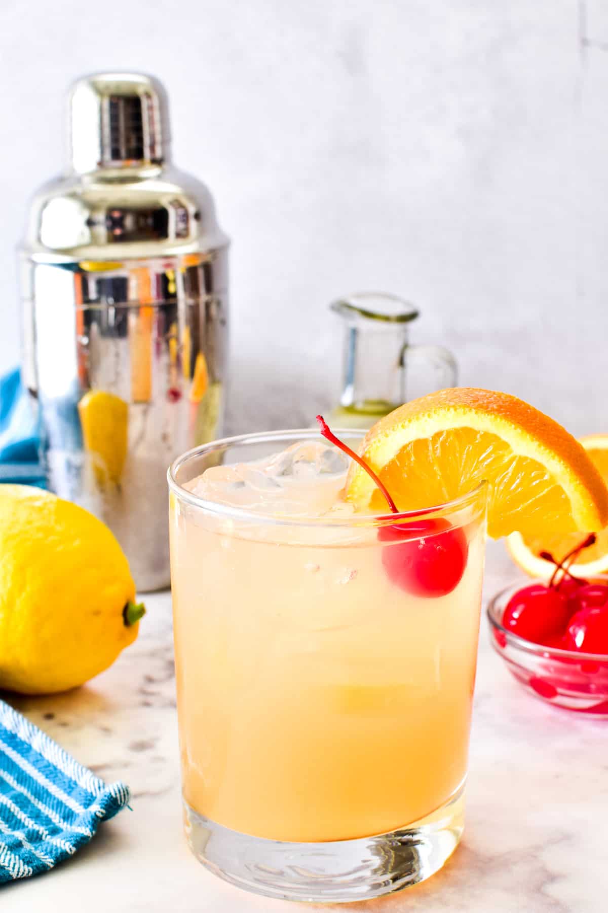 Amaretto Sour garnished with orange and cherry with a cocktail shaker in the background